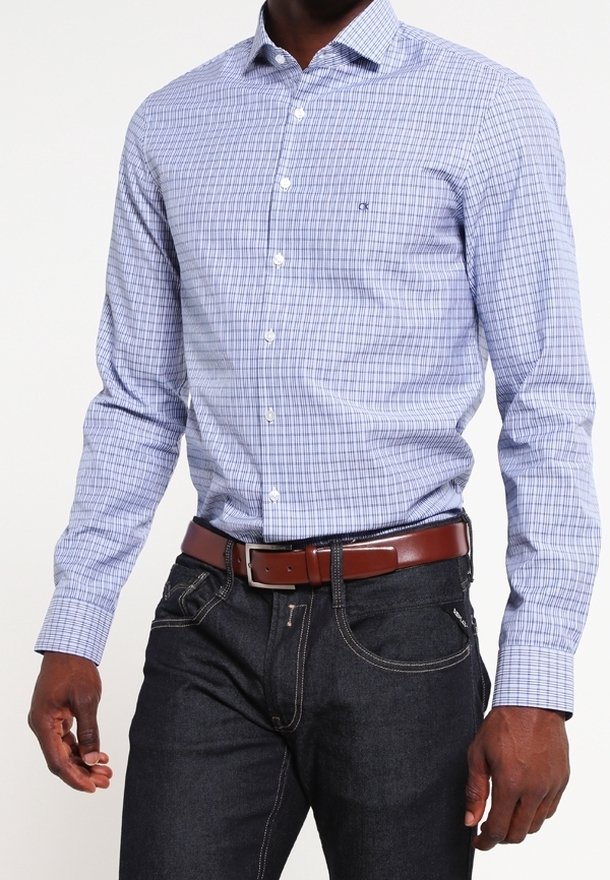 Homme portant une chemise à carreaux bleu clair à manches longues rentrée dans un jean foncé avec une ceinture en cuir marron.