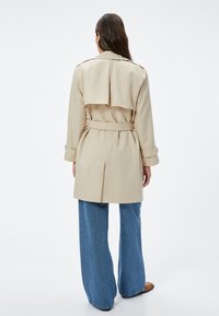 Trench coat beige con spacco posteriore, vita con cintura e spallini sulle spalle. Abbinato a jeans in denim blu a gamba larga. Tessuto di piacevole consistenza.