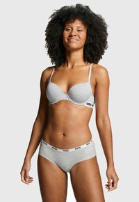 Frau mit lockigem Haar, die einen grauen Puma-BH und passende Unterwäsche trägt, steht selbstbewusst vor einem schlichten hellen Hintergrund.