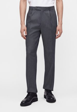 Man draagt donkergrijze nette broek en zwarte leren veterschoenen, staande tegen een effen witte achtergrond.
