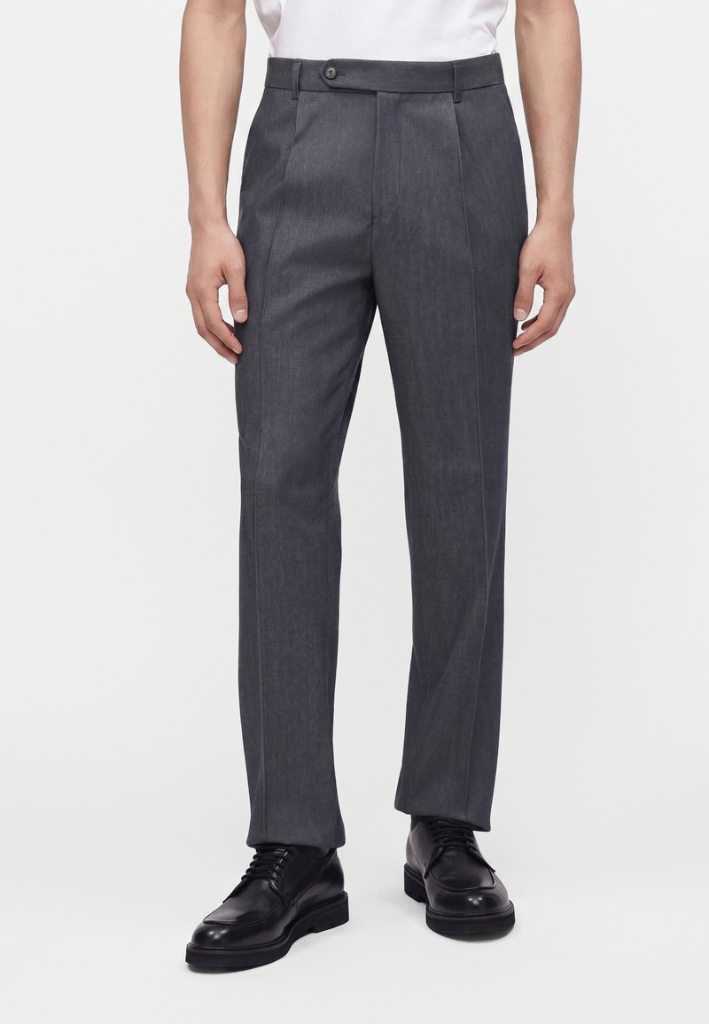 Homme portant un pantalon de costume gris foncé et des chaussures en cuir noires à lacets, debout devant un fond blanc uni.
