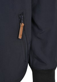 Chaqueta azul marino con una tela texturizada, que cuenta con una cremallera lateral con un tirador de cuero marrón y puños acanalados. Diseño elegante centrado en la utilidad.