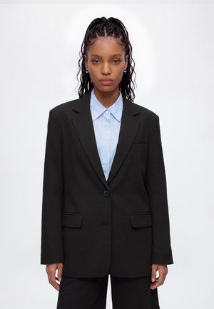 Jeune femme aux cheveux tressés portant une veste de costume noire par-dessus une chemise bleu clair boutonnée, debout sur un fond blanc uni.