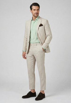 Mann trägt hellbeigen Anzug mit mintgrünem Hemd, braunem Einstecktuch und dunkelbraunen Anzugsschuhen, steht vor einfachem Hintergrund.