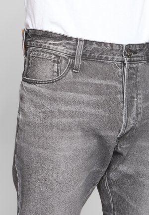 Gros plan d'une personne portant un jean en denim gris délavé avec poches avant, passants de ceinture et une fermeture par bouton noir.