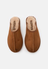 Brune suedine tøfler uten hæl med en fluffy beige innvendig fôr, med en sentral søm og logomerke på innersålen. Myk tekstur gjennom hele.