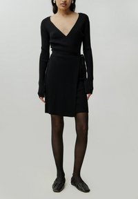Schwarzes geripptes Strickkleid mit tiefem V-Ausschnitt, langen Ärmeln, Wickelstil und einer Bindegürtel in der Taille. Getragen mit schwarzen strukturierten Ballerinas.