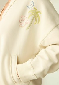 Cremefarvet sweatshirt med V-hals, prydet med broderi af en pink fugl, en gul palme og en blå halvmåne på ærmet.