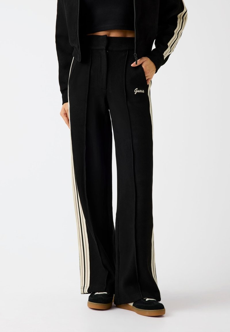Schwarze weit geschnittene Hose mit cremefarbenen Seitenstreifen, hoher Taillenschnitt, weicher Stoff und kleinem gestickten "Guess"-Logo auf der Vorderseite.
