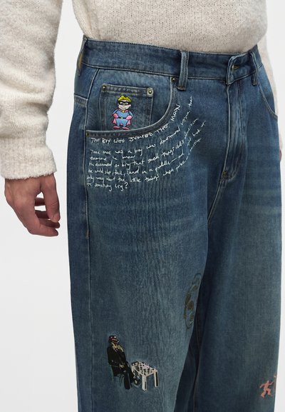 Μπλε τζιν από denim με πολύχρωμες επιδιορθώσεις με σχέδια καρτούν και παίκτη σκακιού και λευκό χειρόγραφο κείμενο κοντά στις μπροστινές τσέπες, φορεμένα με κρεμ πουλόβερ.
