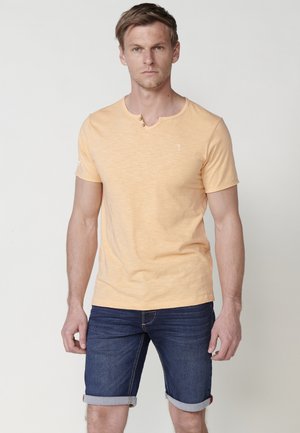 Camiseta de manga corta y cuello redondo en algodón de color naranja claro, con dos botones en el escote y un acabado texturizado. Combinada con pantalones cortos de mezclilla oscura.