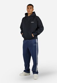 Sort bomulds-hoodie med frontlomme og logo, parret med marineblå træningsbukser med hvide sidestreger og hvide sneakers.