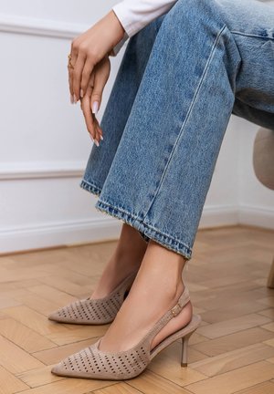 Détail des chaussures montrant des talons beige perforés à bride arrière portés avec un jean court bleu clair sur un sol en bois clair. Les mains reposent sur le genou.