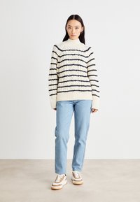 Chunky kremet genser med marineblå horisontale striper, høy hals og ribbestrikkede mansjetter, kombinert med lyseblå jeans og hvite sneakers.