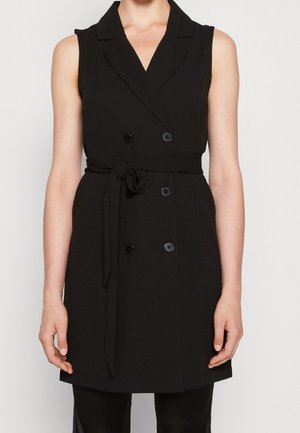 Hihaton musta kaksirivinen liivimekko kauluksella ja vyötärönauhalla, jota käyttää henkilö, jonka kädet lepäävät rentoina sivuilla.