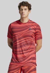 Camiseta atlética roja con patrones de olas y puntos, mangas cortas, cuello redondo y un pequeño logo en el pecho; fabricada con un tejido ligero y transpirable.