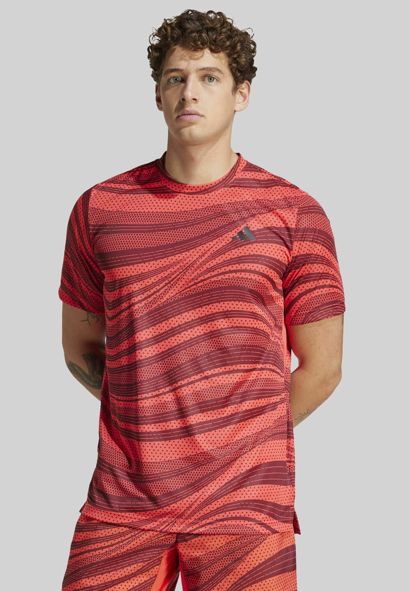 Camiseta atlética roja con patrones de olas y puntos, mangas cortas, cuello redondo y un pequeño logo en el pecho; fabricada con un tejido ligero y transpirable.