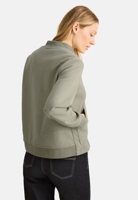 Sweatshirt vert au toucher doux, avec un col subtil et des poches latérales, associé à un jean en denim foncé. Modèle vu de dos.