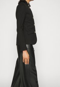 Chaqueta negra ajustada con un cuello alto, con botones plateados y dos bolsillos frontales. Combinada con pantalones de cuero negros de talle alto.
