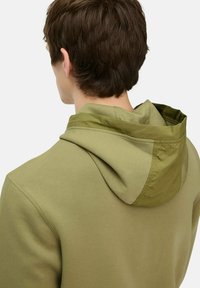 Olijfgroene hoodie met een contrasterende lichtgewicht capuchon, een zachte textuur en een relaxed fit. Naaddetails zichtbaar op de schouders.