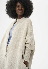 Beżowy sweter z dzianiny z wysokim kołnierzem, luźnym krojem i przednim zamkiem. Posiada teksturowany wzór dzianiny oraz rozcięte rękawy dla swobody ruchów.