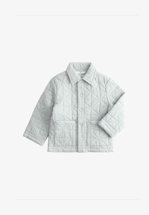 Ung dreng iført let grå quiltet jakke, løse blå jeans, marineblå sneakers, der holder lyse lilla solbriller, stående mod hvid baggrund.