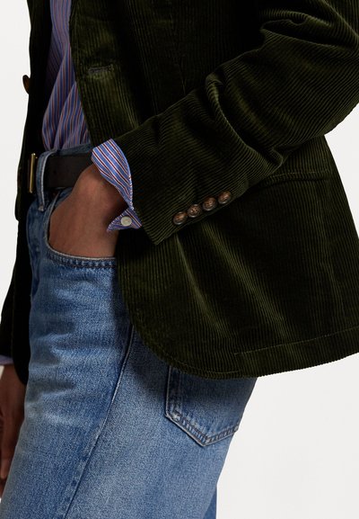 Dark green corduroy blazer with vertical ribs, featuring brass buttons and a classic collar. Worn over a striped shirt and blue jeans.