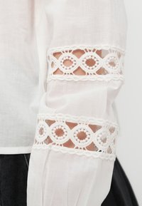 Λευκό μακρυμάνικο με κυκλικές δαντελωτές ανοίγματα που αποκαλύπτουν το δέρμα στο πάνω μέρος του βραχίονα, φορεμένο με σκούρο παντελόνι πάνω σε απλό φόντο.