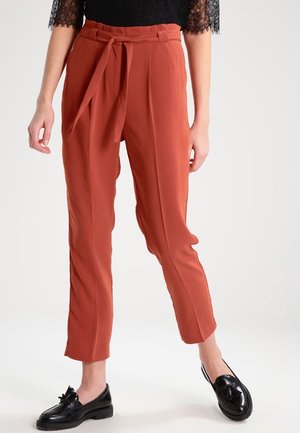 Pantalon taille haute fuselé couleur rouille avec lien à la taille, porté avec un haut en dentelle noire et des mocassins noirs à pompons sur fond clair.