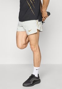 Light grey running shorts with golden Nike logo and accents, paired with black athletic shoes and white socks. Minimalist design, cut above knee.