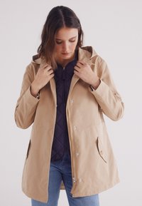 Impermeabile beige con cappuccio, chiusure a scatto e tasche laterali. Indossato sopra uno strato interno trapuntato scuro, esposto su uno sfondo neutro.