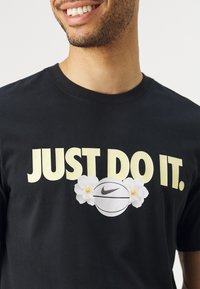 Svart bomull T-skjorte med "JUST DO IT." i stor gul skrift, med en basketballgrafikk og hvite blomsteraksenter.