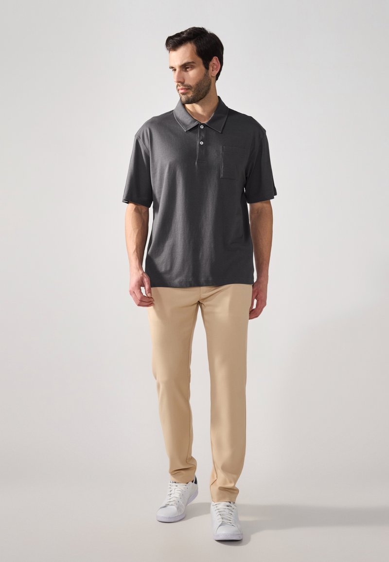 Polo grigio con una sola tasca sul petto, abbinato a pantaloni beige chiaro e sneakers bianche. La camicia ha un colletto classico e maniche corte.