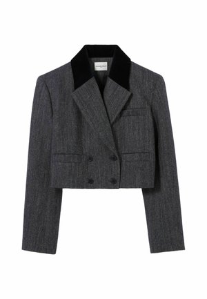Jeune femme portant un blazer gris court et une jupe, tenant un sac à main rouge, avec des chaussettes rouges et des chaussures marron foncé à lacets.