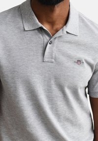 Homme portant un polo gris clair avec un petit logo en forme de bouclier brodé rouge, blanc et bleu sur la poitrine gauche.