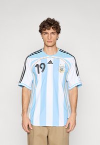 Η φανέλα της εθνικής ομάδας ποδοσφαίρου της Αργεντινής με κάθετες ριγες σε γαλάζιο και λευκό. Μαύρες ρίγες Adidas στα μανίκια και λογότυπο· αριθμός παίκτη 19.