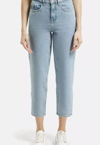 Kobieta w jasnoniebieskich, skrojonnych dżinsach o prostym kroju i białych trampkach, stoi na jednolitym tle, z rękami luzno opuszczonymi po bokach.