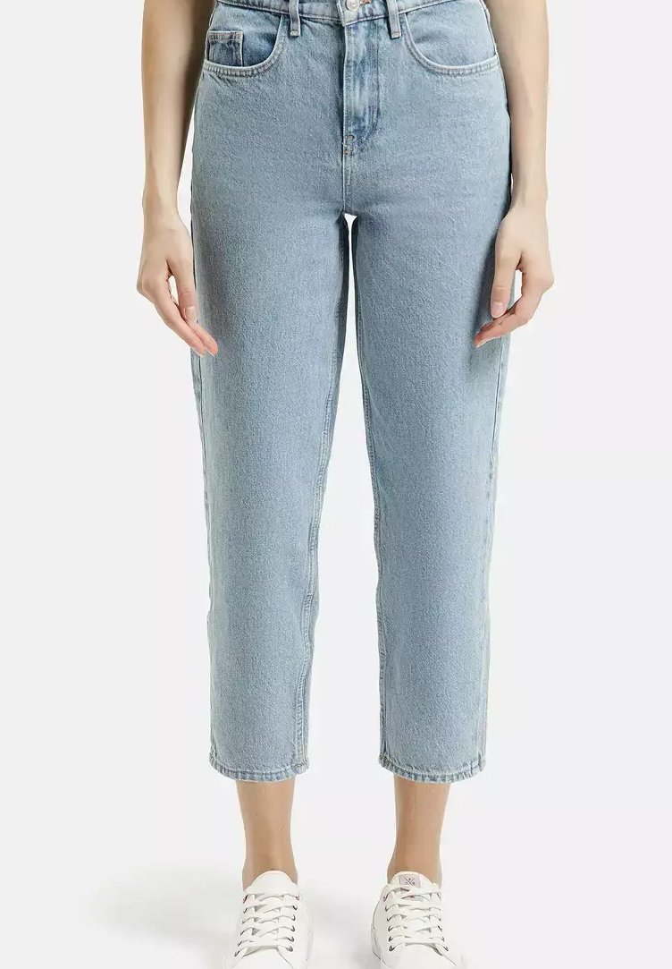 Kobieta w jasnoniebieskich, skrojonnych dżinsach o prostym kroju i białych trampkach, stoi na jednolitym tle, z rękami luzno opuszczonymi po bokach.