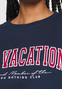 Person klädd i en marinblå sweatshirt med röd och vit text som står "VACATION" och mindre text "Stolt medlem av DO NOTHING CLUB."