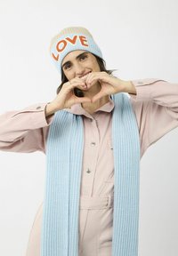 Gebreide beanie in beige en lichtblauw met "LOVE" in oranje; lichtblauwe geribbelde sjaal; roze blouse. Handen die een hartvorm vormen.