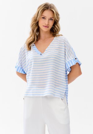 Kobieta o falowanych blond włosach, ubrana w luźną, krótką bluzkę w jasnoniebiesko-białe paski z dekoltem w serek i białe spodnie, stojąca z rękami za plecami.