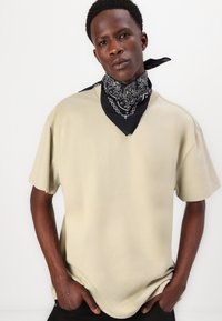 Chemise beige à manches courtes, coupe relaxée, en tissu doux. Bandana noir autour du cou avec des motifs blancs complexes.