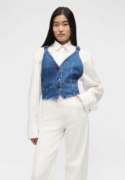 Γυναίκα που φοράει λευκό πουκάμισο και παντελόνι με μπλε τζιν γιλέκο με κουμπιά, στέκεται με το ένα χέρι στην τσέπη μπροστά σε απλό φόντο.
