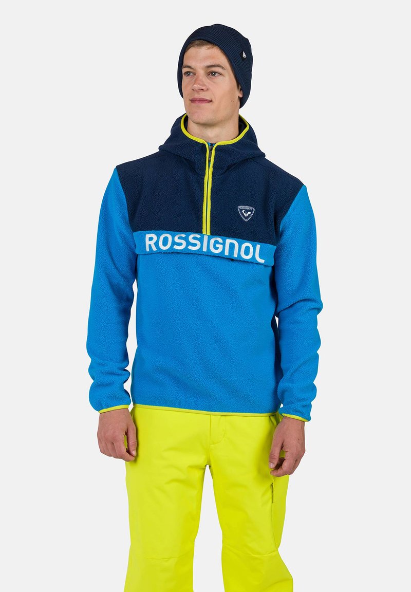 Sininen ja tummansininen fleece-pullove, jossa on keltainen vetoketju. Siinä on logo-patch ja "ROSSIGNOL"-brändäys rinnassa valkoisella fontilla.