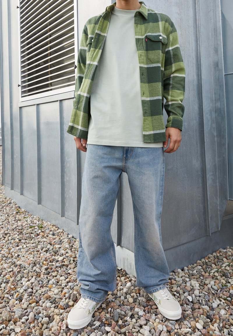 Chemise en flanelle à carreaux verts et gris portée sur un t-shirt vert clair, associée à un jean bleu délavé et des baskets blanches avec des accents brodés.