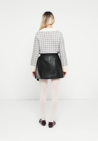 Donna con lunghi capelli biondi che indossa un maglione a scacchi, una gonna in pelle nera, collant bianchi e scarpe basse rosso scuro, in piedi contro un muro bianco.