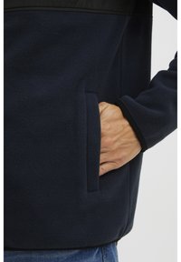 Giacca in pile blu marina con tasca laterale, pannello nero sopra il petto e polsini neri. Morbida al tatto con una vestibilità comoda.