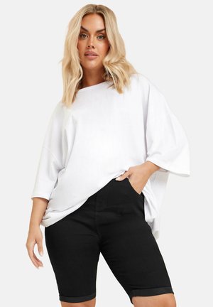Camiseta blanca de gran tamaño con mangas cortas, combinada con pantalones cortos de mezclilla negros. Los pantalones cortos tienen dobladillos enrollados y bolsillos laterales.