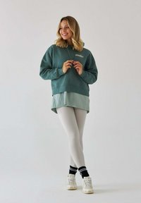 Zielona bluza z kapturem z logo, nałożona na jasną zieloną koszulę, w połączeniu z białymi legginsami i teksturowanymi snekerami. Prosty design, luźny krój.