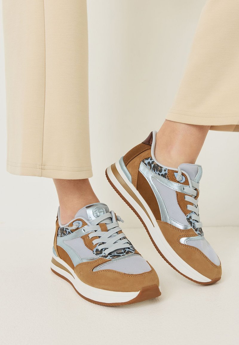 Pieds portant un pantalon beige et des baskets stylées marron, argentées et à imprimé léopard avec des semelles blanches sur un fond clair.
